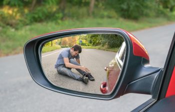 A man sits on the road holding his ankle, reflected in a car's side mirror on a country road.