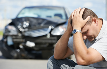 A man sits on the ground holding his head in distress; a damaged car is in the background.