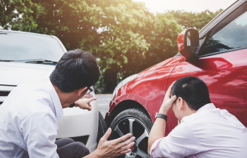 Two men assess minor damage between a red and a white car on a street. One man points at the cars while the other holds his head with one hand. Trees are visible in the background.