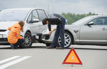 Two people inspect minor damage on the front of two cars after a collision on a road. An emergency triangle is placed behind the vehicles.