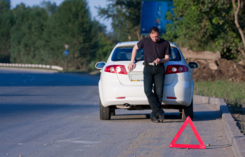 A man leans against a white car parked on the roadside, using a smartphone. A red warning triangle is placed on the ground behind the vehicle. Trees line the background.