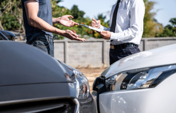 Two people stand in front of two cars involved in an accident. One person is holding a clipboard and pen, while the other gestures with their hands.